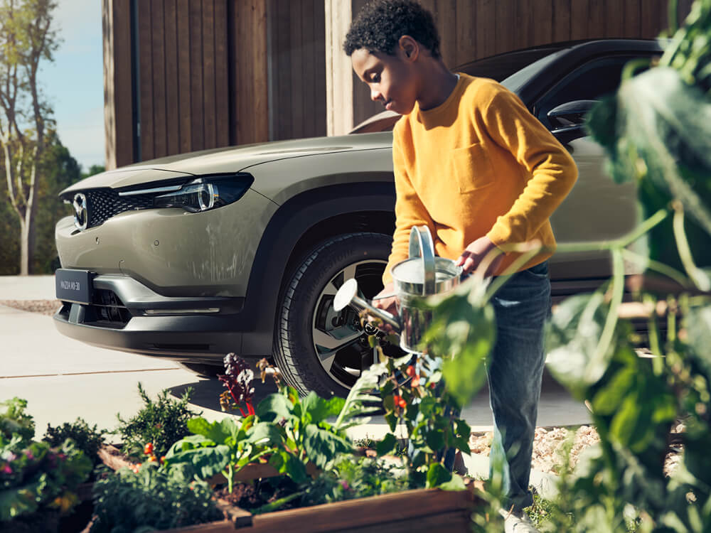 Young boy watering his garden, with a Mazda vehicle in the background