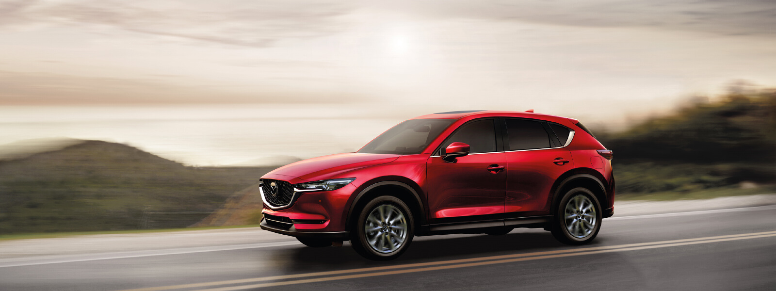 Soul Red Crystal Mazda SUV drives along rural highway with brown hill country blurred in the background. 