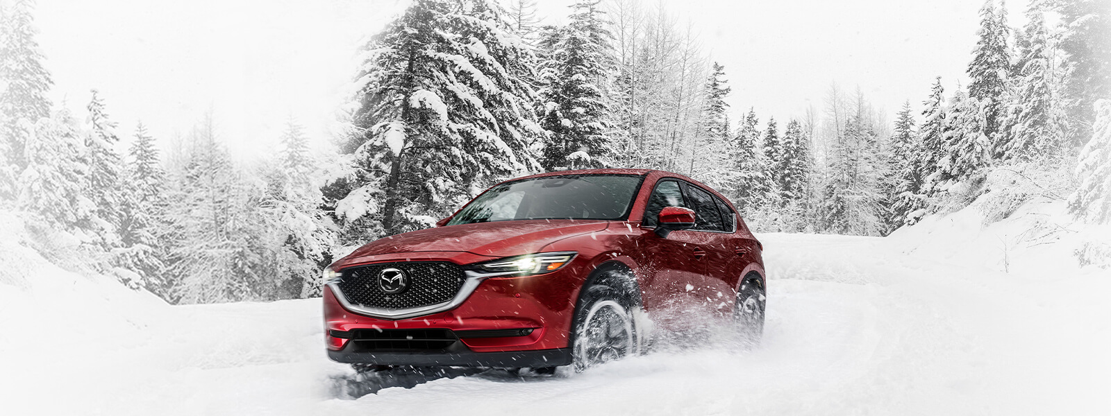 Soul Red Crystal Mazda SUV with headlights on drives past snow-covered conifers as it negotiates a curve in deep snow on a rural road. 