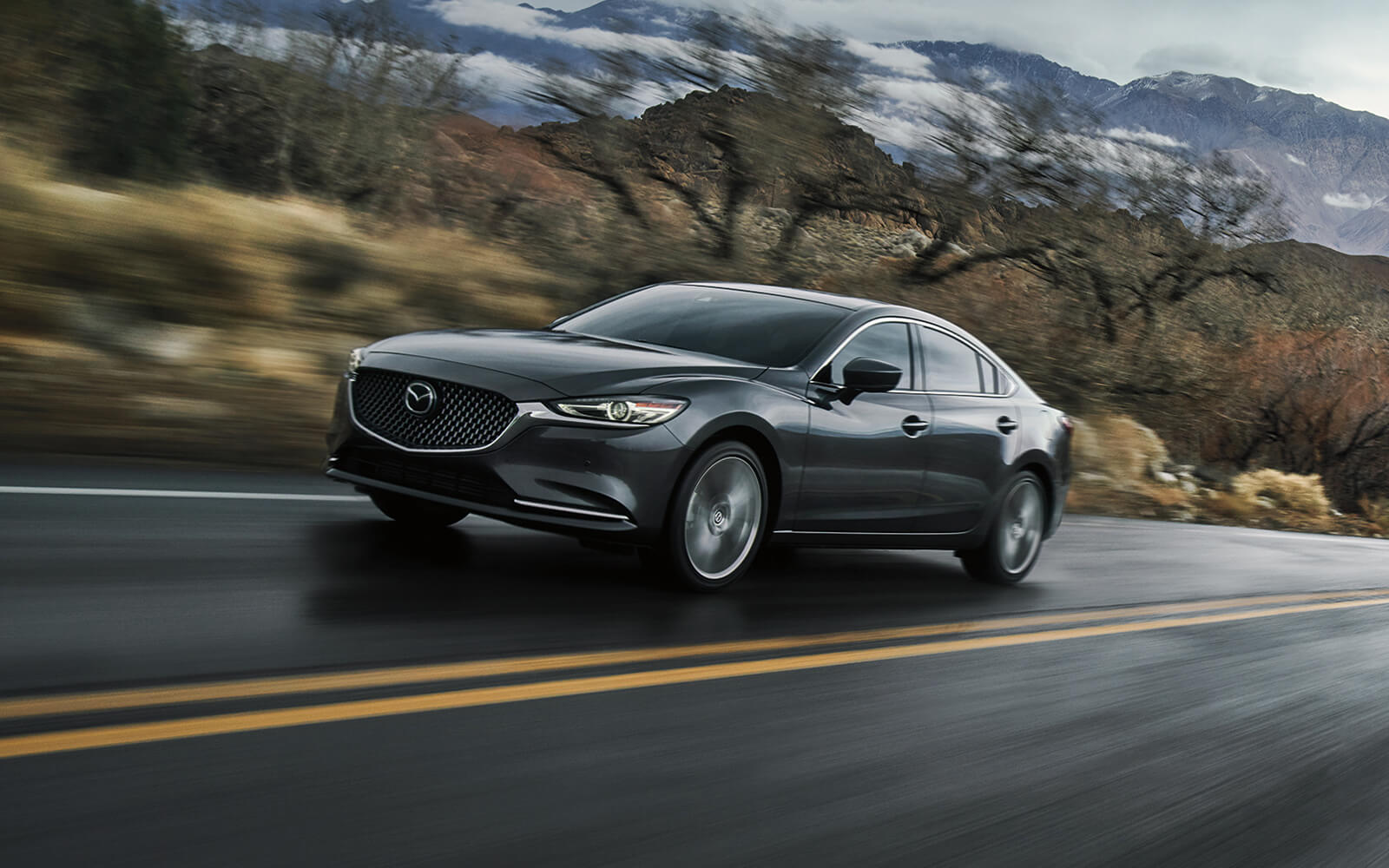 Grey Mazda6 ascending highway hill with blurred trees and hills in background 