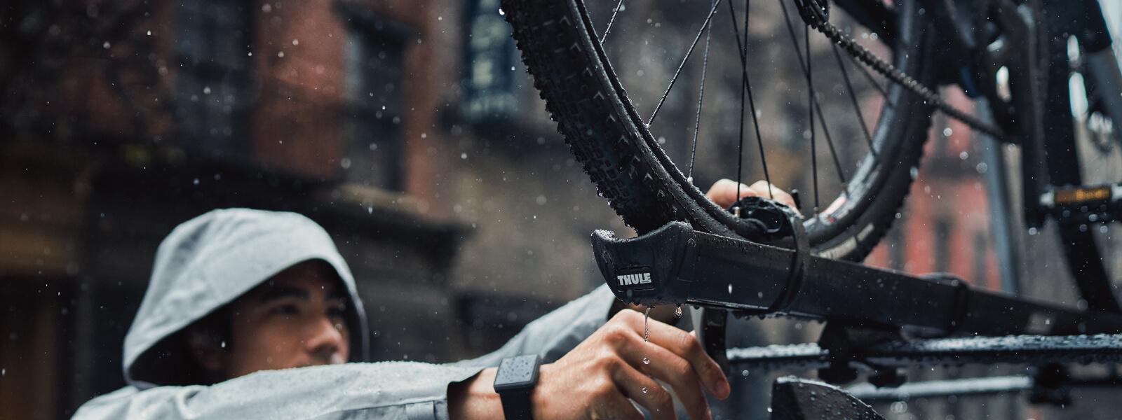 Un jeune homme portant un imperméable gris avec le capuchon sur la tête ajuste la sangle qui retient la roue du vélo au porte-bagages