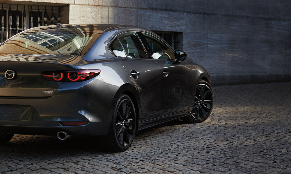 Rear passenger-side tail light view of grey Mazda3 sedan turning on urban cobblestone path. 