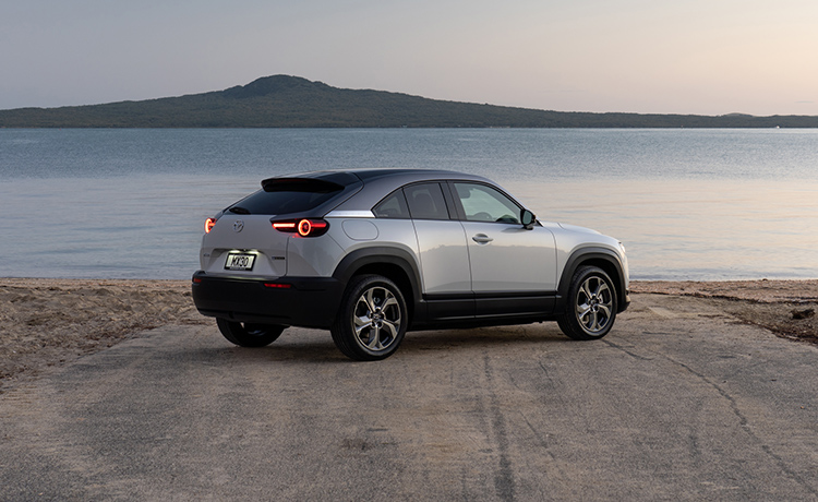 Silver Mazda MX-30 EV viewed from behind rear passenger side on beach with coastline on horizon.