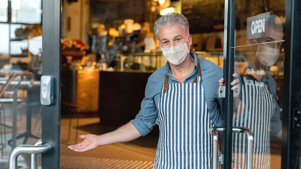 A man wearing a face mask and apron holds open the door to a store, welcoming the viewer inside with one arm.