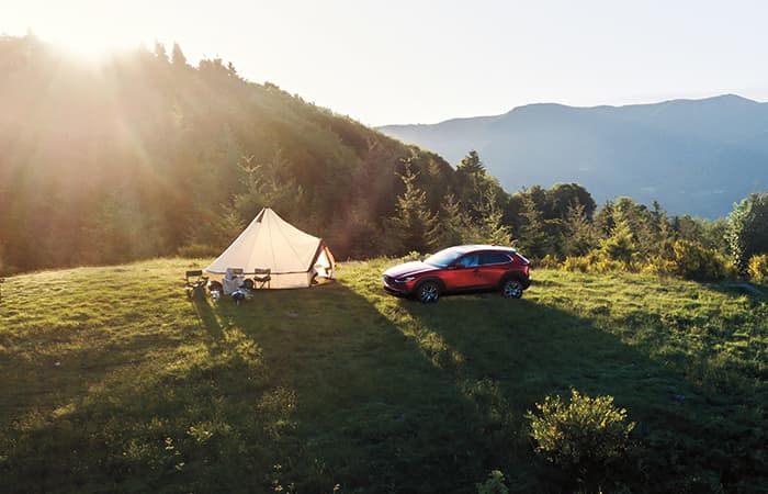 A red Mazda vehicle parked next to a tent in a forested area.