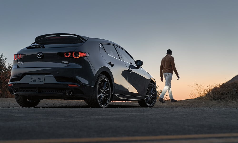 Une Mazda3 Sport noir de jais mica stationnée sur le bord de l’autoroute, alors que le conducteur se dirige vers un sentier, avec un coucher de soleil en toile de fond