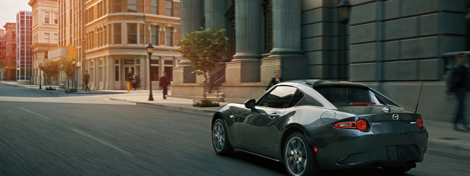 Polymetal gray MX-5 2-seat convertible driving down urban street past pedestrians blurred in background.