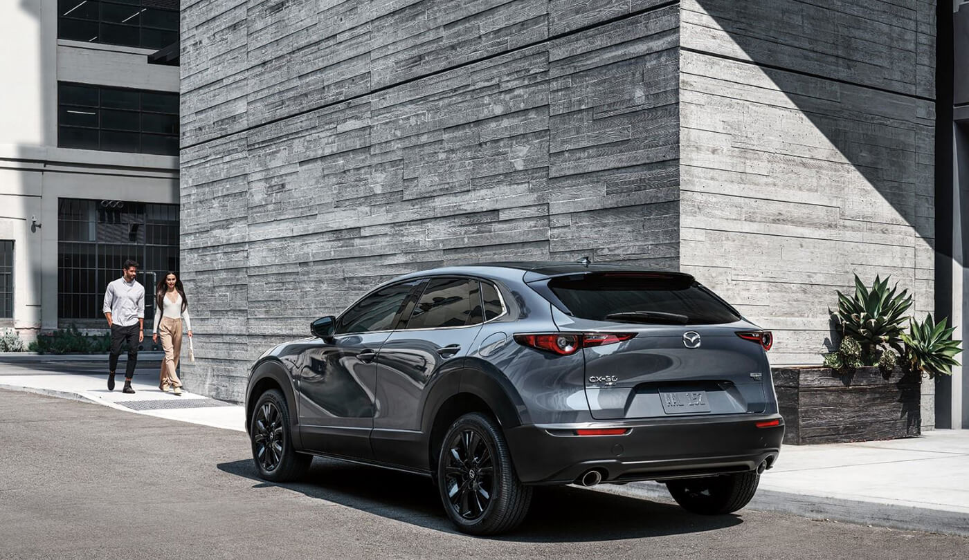 Mica Blue Mazda CX-30 parked in sunshine next to modern building as young professional couple approach on sidewalk.