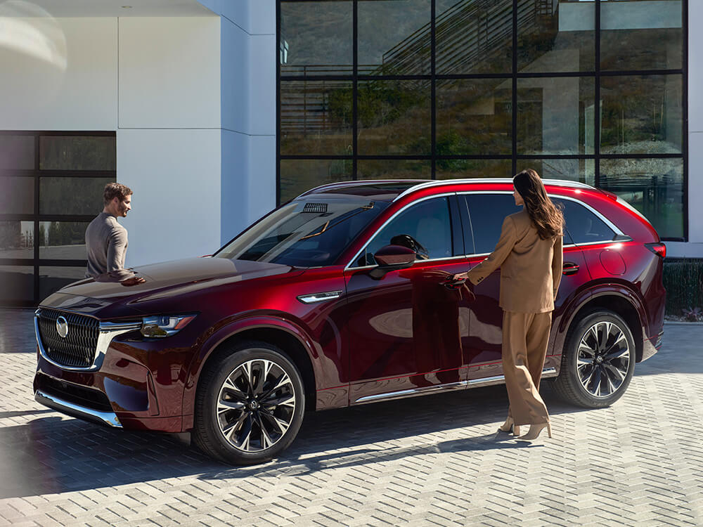 Artisan Red CX-90 parked in front of stylish building. A woman is opening the driver’s side door, a man is going to the passenger side. 