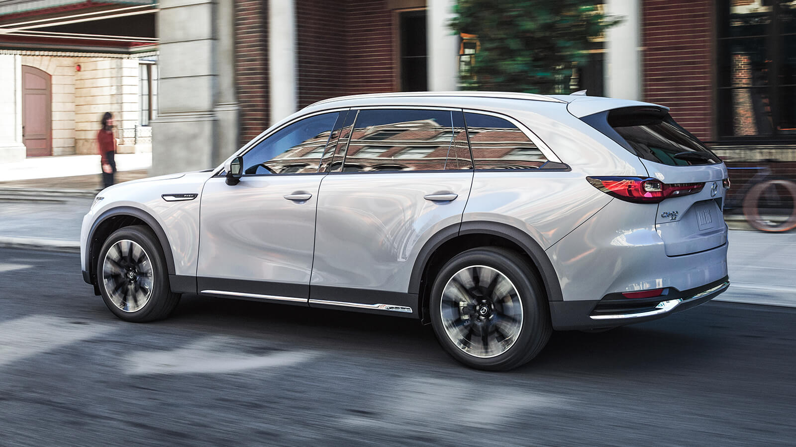 Un VÉHR CX-90 blanc arctique dépasse un piéton devant un bâtiment en briques rouges dans une rue urbaine. 