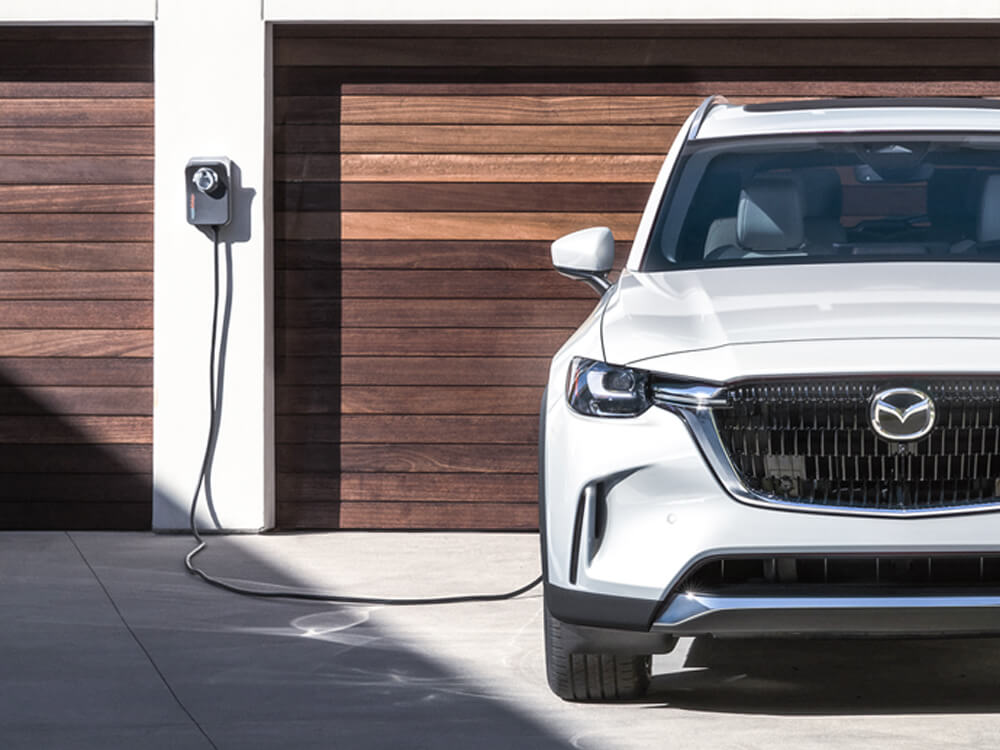 Un VÉHR CX-90 blanc arctique, branché et en mode recharge, garé devant un garage double avec des portes de garage à lattes de bois.  