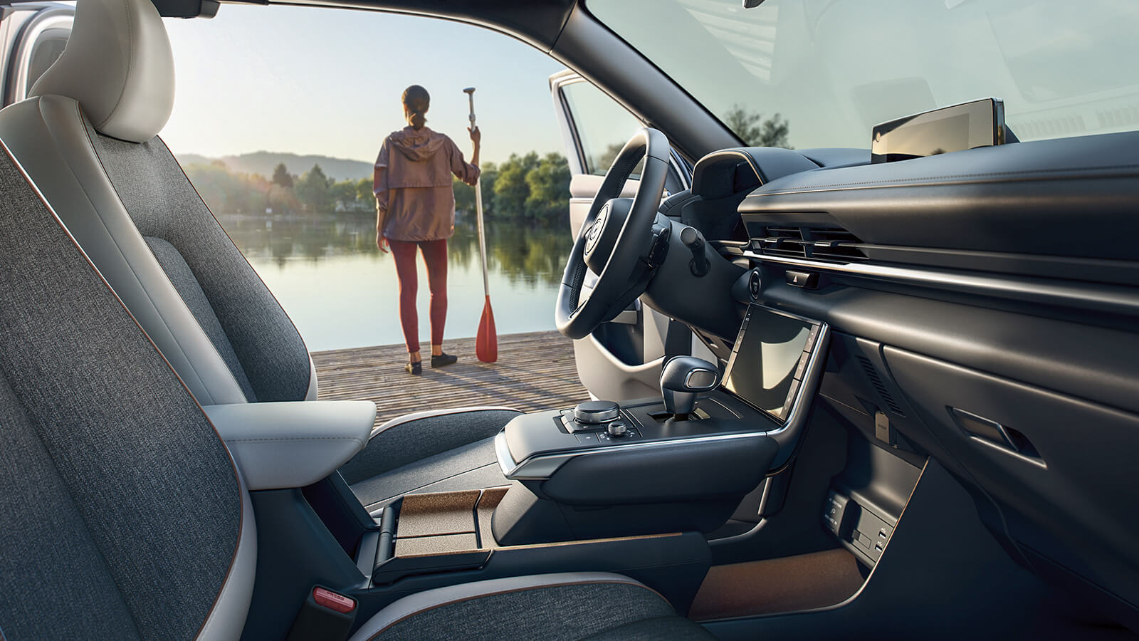 Shot from the front passenger seat looking out of the open driver’s side door, a woman stands on a wooden dock at sunrise looking over a lake with a paddle in her hand.  