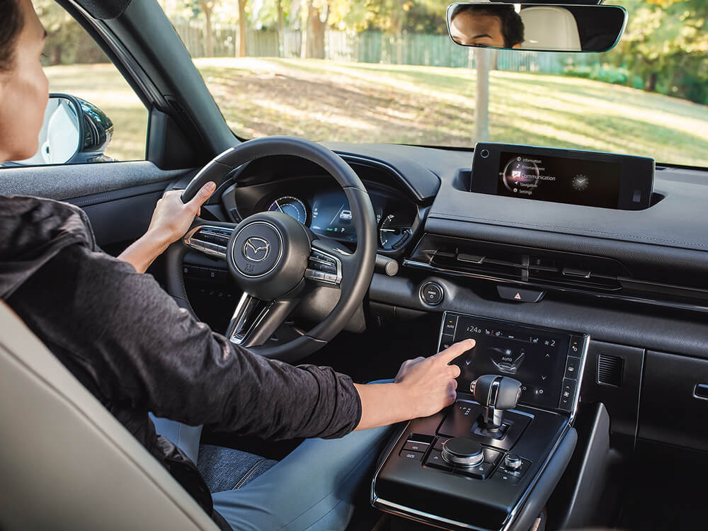 Shot from back seat shows driver inside the MX-30 tapping the full colour display’s touch screen in the centre console. 