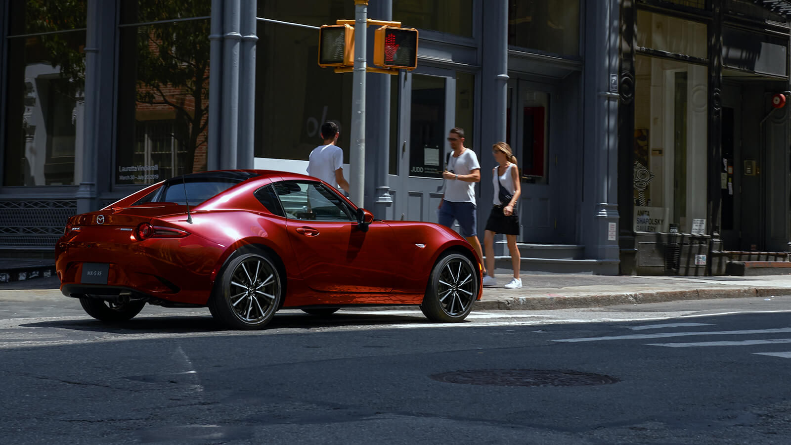 Une MX-5 RF rouge vibrant cristal métallisé arrêtée à un feu rouge dans une rue principale; des piétons passent en arrière-plan.