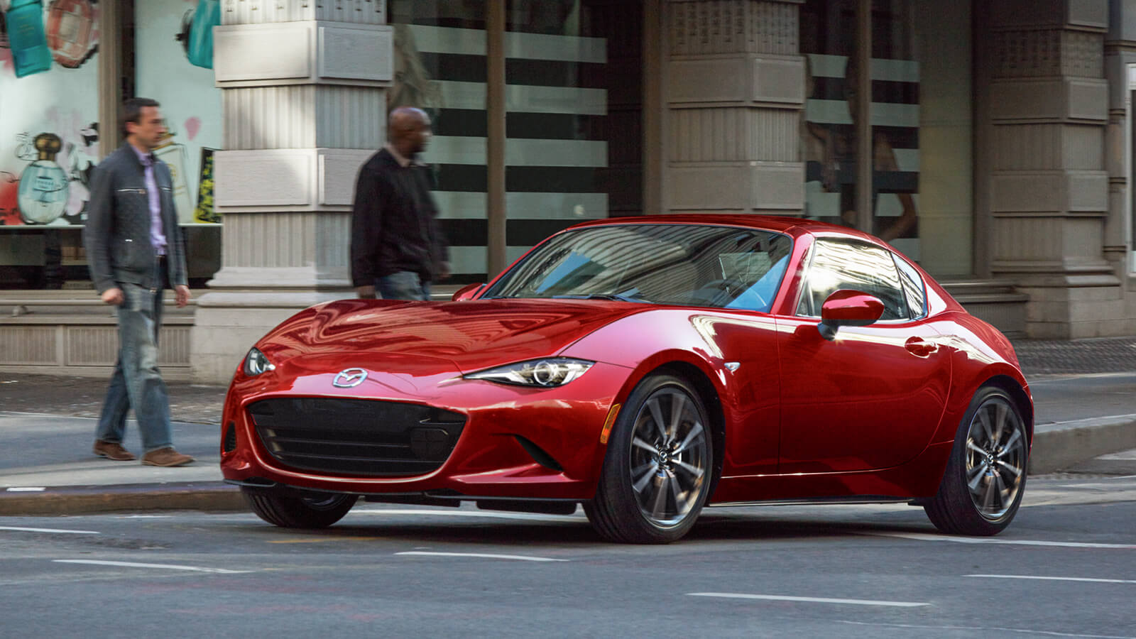 Une MX-5 RF rouge vibrant cristal métallisé avec le toit fermé s’éloigne d’un passage pour piétons; des gens attendent pour traverser en arrière-plan.