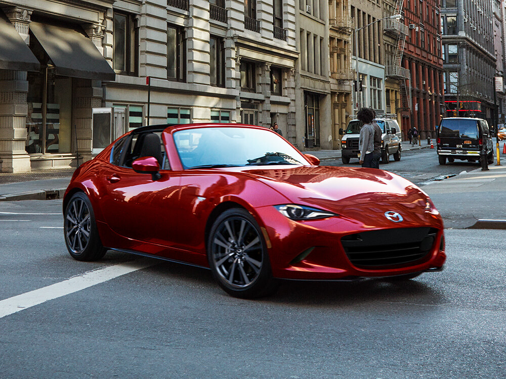 Une MX-5 RF rouge vibrant cristal métallisé reflète les bâtiments environnants en effectuant un virage à gauche dans une ville animée.