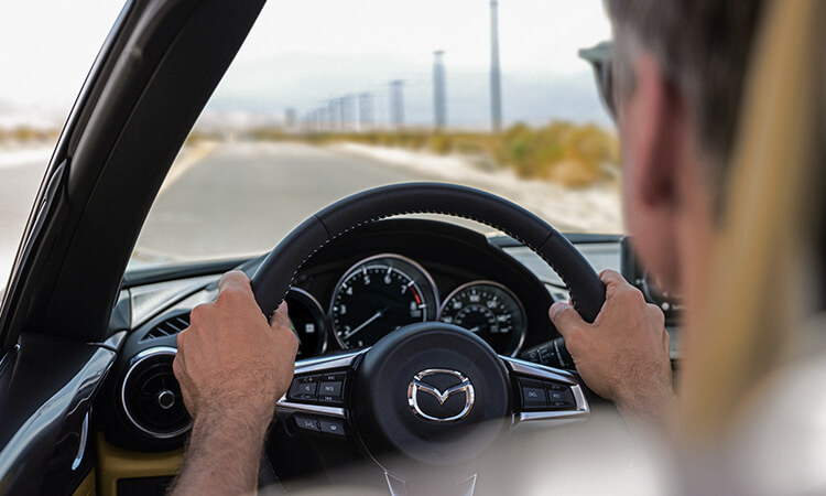 Vue arrière du conducteur d’une MX-5 ST; les mains sur le volant, il regarde une route dégagée bordée de poteaux électriques. 