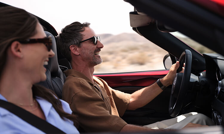 Vue du côté passager à l’intérieur d’une MX-5; le passager et le conducteur rient en roulant avec le toit baissé.