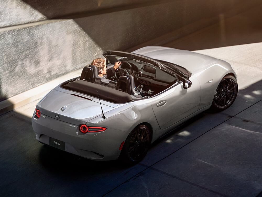 Une MX-5 gris aéro métallisé traverse un rayon de soleil matinal en sortant d’un stationnement souterrain.