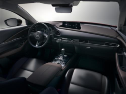 Dans un environnement de studio transparent, vue de l’intérieur d’un CX-30 du côté passager vers l’avant et vers le bas sur les sièges avant, le volant, le tableau de bord, la console et l’écran.