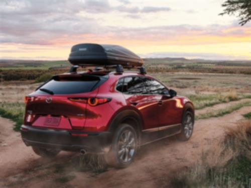 Plan de ¾ arrière d’un CX-30 rouge vibrant cristal métallisé avec coffre de toit, soulevant la poussière en s’éloignant sur un chemin de terre dans un environnement rural.