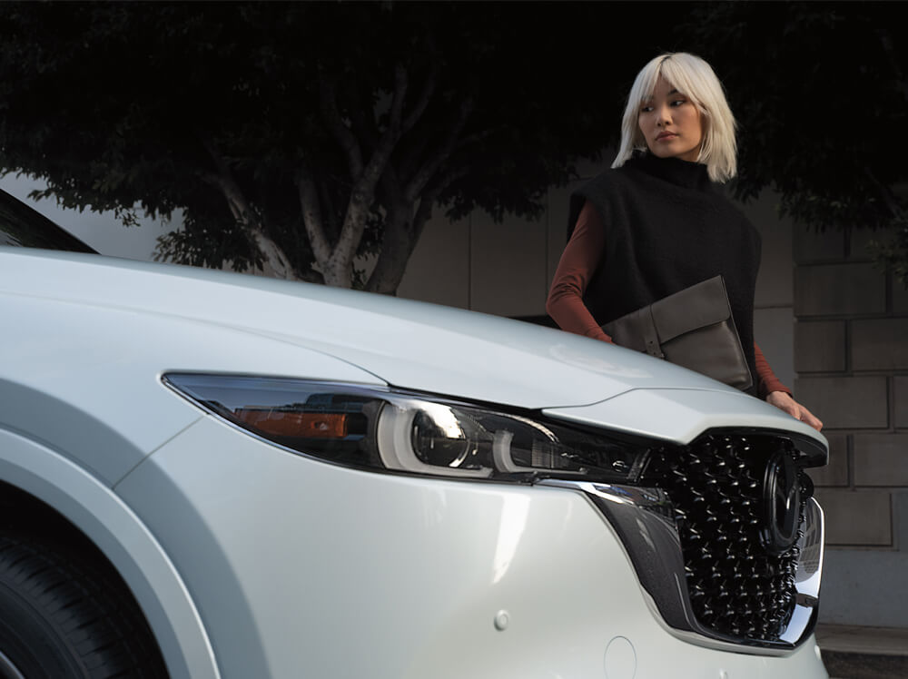 Hood and grille of Rhodium White Metallic CX-5 in low foreground, stylish woman looks back at windshield.