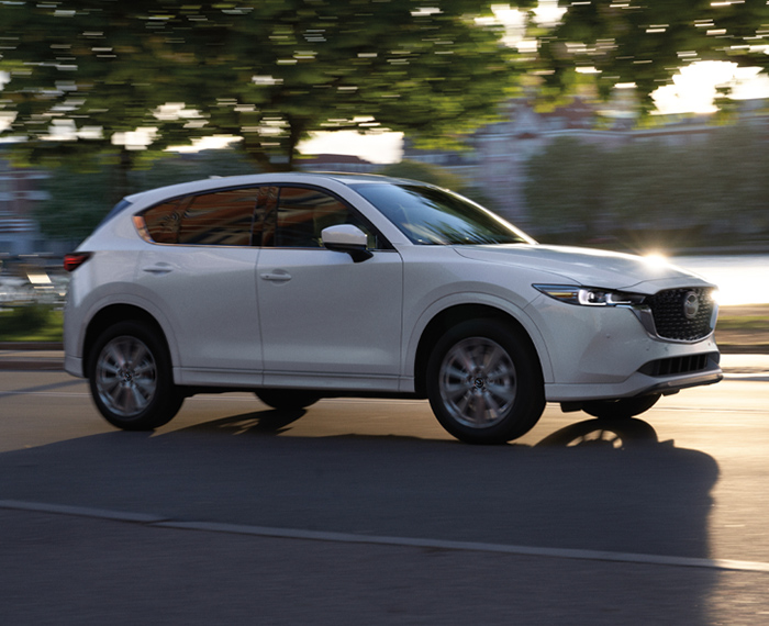 Rhodium white CX-5 driving profile shot, stately building and trees motion blurred in background. 