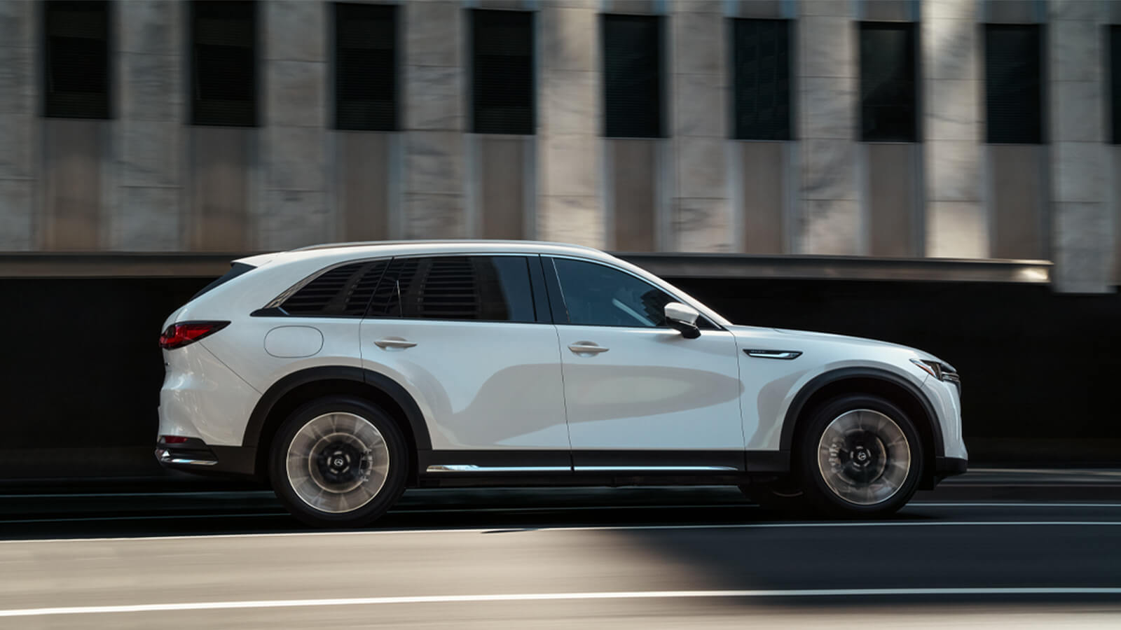 Profile view of Arctic White CX-90 PHEV driving past minimalist, modern concrete building. 