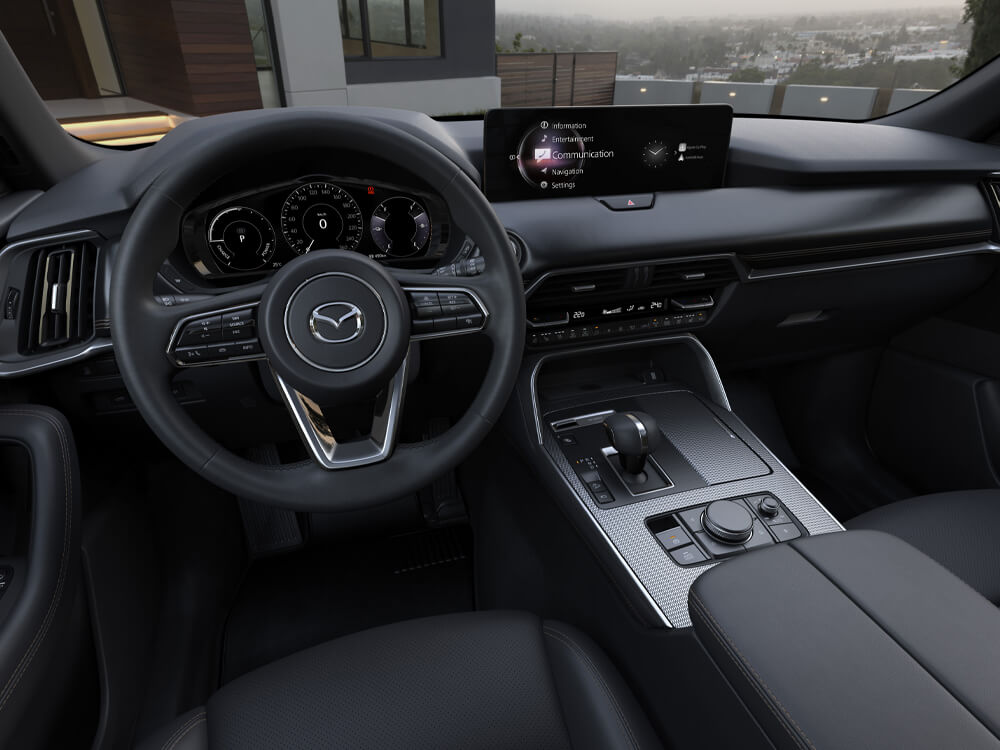 Driver’s seat view of steering wheel, dash, console in CX-90 parked in front of a stylish house. 