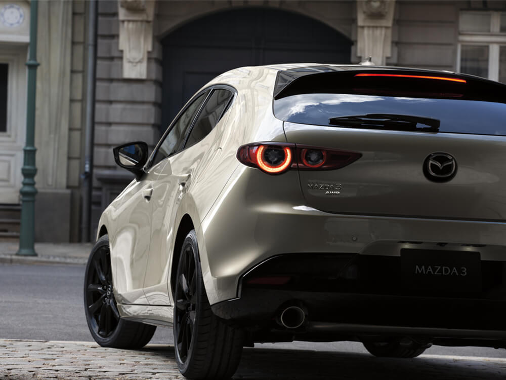 Shot low back Suna Edition Mazda3 Sport on cobblestone street. Close-up of Mazda3 & AWD badge & Mazda logo on rear hatch door.