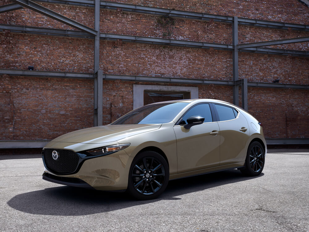 Suna Edition Mazda3 Sport parked in the sun, profile. Raw brick and steel girder building in background. 