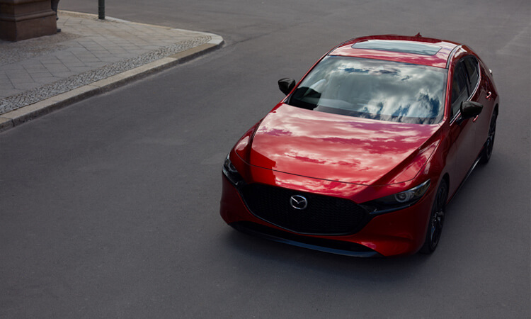 Above ¾ shot of Mazda3 Sport driving down city street, sky and clouds reflected in hood and windshield. 